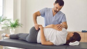 Male osteopath in blue scrubs performing a manual hip and back adjustment on a female patient lying on a treatment table in a bright clinic.