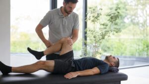 Male osteopath in a grey polo shirt performing a knee mobilization stretch on a man lying on a massage table in a bright, modern clinic.
